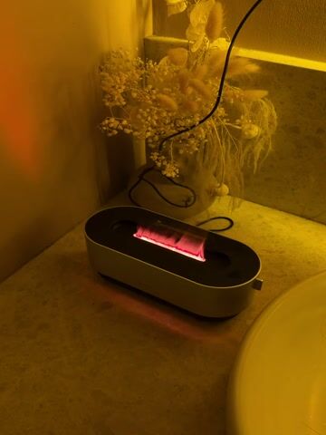 The video captures a close-up shot of a small, oval-shaped electronic device with a simulated flame effect, placed on a light-colored countertop. The device has a dark top surface and a lighter, possibly off-white or beige, base. Inside a rectangular opening on its top, a reddish-pink light flickers, creating the illusion of small flames. The light from these simulated flames reflects onto the countertop beneath the device.

Behind the device, a vase holds a bouquet of dried flowers, predominantly light-colored, feathery elements that resemble pampas grass and small white flowers, possibly baby's breath. A black power cord is visible, extending from the device upwards and out of the frame. The entire scene is bathed in a warm, yellowish artificial light, which casts a soft glow over the objects and the background wall. To the right, a portion of a white sink or basin is visible, suggesting the setting is a bathroom or vanity area.

A small label or card with some text is visible inside the device, near the flickering "flames," but the text is too small and indistinct to read clearly. The video is static, focusing solely on the operating device and its immediate surroundings.