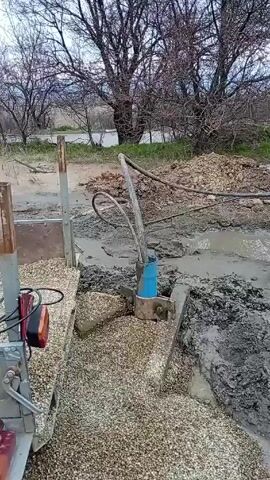 The video shows a muddy construction site. Water is being pumped out of a hole surrounded by gravel and mud. There are pipes and other equipment scattered around the site. In the background, there are bare trees and an overcast sky. A pile of mud is also present, along with an old barrel and a tire. The environment seems rural or undeveloped.