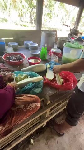 The video shows two people preparing food at a rustic wooden table outdoors. The person on the left, wearing a maroon long-sleeved shirt and an orange and brown patterned skirt, is peeling a light green vegetable with a knife. The vegetable is placed in a teal plastic bag. To their right, the person on the right, wearing a blue and white striped shirt and dark shorts, is also cutting a similar light green vegetable, which is inside a red plastic bag. There are several whole, elongated light green vegetables on the table. The table is cluttered with various items, including stacks of white plates, red bowls containing food, a large metal pot, two green bottles, a clear plastic container with green liquid, and a blue container. The environment appears to be a tropical outdoor setting with trees visible in the background and a wooden structure overhead.