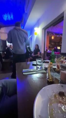 The video shows a man and a woman dancing in a restaurant. The man is wearing a light gray shirt and dark pants, and the woman is wearing a black dress. They are holding each other close. The restaurant has a casual atmosphere, and there are other people sitting at tables in the background. There are also empty plates and glasses on the table in the foreground. The lighting changes color throughout the video.