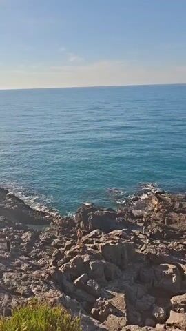 The video shows a rocky coastline meeting the ocean on a clear, sunny day. The camera looks out from a slightly elevated rocky area towards the calm, blue ocean stretching to the horizon under a clear blue sky.  The rocky foreground consists of large, dark-colored boulders and smaller rocks. In the distance, a person stands on the rocks. Small waves gently lap against the rocky shore. The overall setting suggests a serene coastal environment.