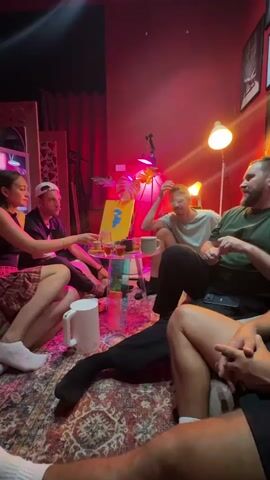 A group of five young adults are gathered in a room with red lighting. A woman on the left is wearing a plaid skirt and white socks, sitting with her legs crossed. Next to her is a man in a black shirt and a white baseball cap, also seated. A third person, wearing a white t-shirt, is sitting on the floor in the center, facing the group and holding a small glass. To the right, a man in a green t-shirt is gesturing with his hands while talking, and another person, mostly out of frame, is seated in the foreground with their legs visible. In the center of the group, there is a low table with several small glasses containing liquid, a white electric kettle, and a framed piece of art with blue and yellow shapes. The room has a patterned rug on the floor and curtains in the background. The overall atmosphere appears casual and social, possibly during a gathering or conversation.