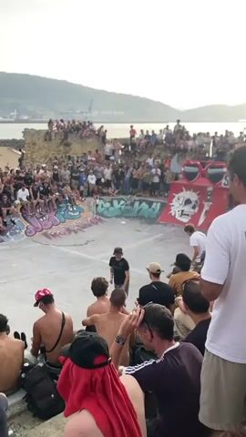 A crowded outdoor skatepark is the setting for a skateboarding event. The scene is viewed from behind a group of spectators seated on a tiered embankment. The sun is setting, casting a warm, hazy glow over the sky and the distant water. In the foreground, several people are seated, with their backs to the camera, watching the event. The middle ground features a skate ramp with a large graphic of sunglasses and a skull. A skateboarder in a white t-shirt and black shorts is performing a trick on the ramp. The background shows a body of water, mountains, and a harbor with cranes. The skatepark itself is concrete with graffiti art on the walls of the bowl. The overall atmosphere appears to be energetic and festive, with a large crowd gathered to watch the skateboarding performance.