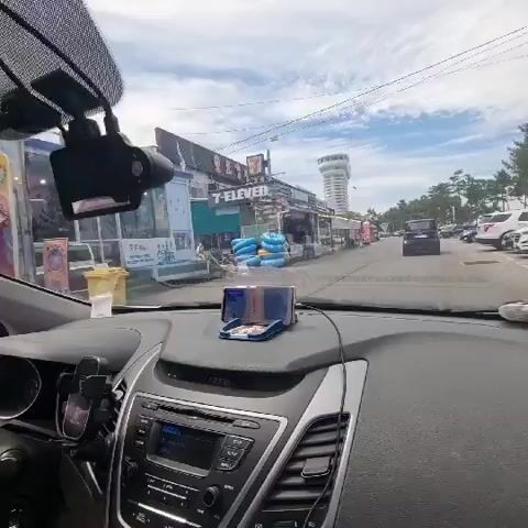 The video shows a view from inside a car looking out the windshield. The car is driving on a street lined with shops and businesses, many of which appear to be restaurants or food stalls. The shops have colorful signs and awnings. There are several cars parked along the side of the road and some people walking in front of the shops. In the distance, a tall, white tower-like structure is visible. The sky is partly cloudy. A smartphone is mounted on the dashboard in front of the driver, displaying something. A dashcam is visible mounted on the rearview mirror.