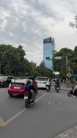 The video shows a busy city street with moderate traffic. A tall, modern skyscraper with a blue glass facade dominates the background. Several cars and motorcycles are on the road, some stopped at a traffic light. The traffic light is cycling through green, yellow, and red. Trees line the sides of the street, and the sky is overcast with clouds. There are traffic signs visible, including a speed limit sign. The scene is dynamic, with vehicles and people moving.