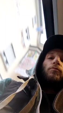 A man with a beard and a black baseball cap sits by a window on a moving vehicle. Buildings can be seen through the window. The man wears a dark hoodie and a jacket. The video is shaky.
