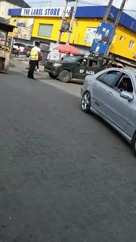 The video shows an intersection on a street with cars, motorcycles, pedestrians, and buildings. A man in a yellow vest appears to be directing traffic. Cars including a silver Mercedes-Benz, a silver Toyota Corolla, and a blue van are present. There is a truck with "OP MESA" written on it. A building labeled "THE LABEL STORE" is visible, painted in yellow and blue. There is also an advertisement poster on the side of the building. The street is paved with interlocking stones, and there is litter visible on the ground.