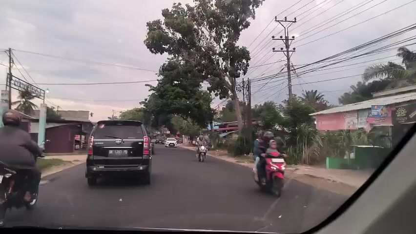 The video shows a drive down a street in a small town on an overcast day. The camera is inside a car, looking out the front windshield at the road ahead.  Several cars and motorcycles are on the road. Buildings and trees line the sides of the road, and overhead are power lines.