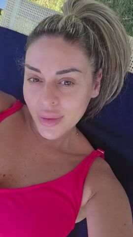 The media file is a close-up selfie video of a woman lying down outdoors. She is wearing a bright pink, low-cut swimsuit. Her hair is pulled back into a high ponytail, and she has light makeup on, with her eyes and lips being the most prominent features. She is looking directly at the camera with a slight smile.

The setting appears to be a sunny day, as indicated by the natural light and the background which shows green grass and what looks like a woven lounge chair. The overall impression is one of relaxation and leisure, possibly at a resort or a private garden. There is no visible text in the video. The woman's expression suggests a calm and contented mood. Given the context of Reggio nell'Emilia, Italy, this scene could be from a vacation or a day spent enjoying good weather in a pleasant outdoor environment. The lighting suggests it is daytime, likely midday or afternoon.