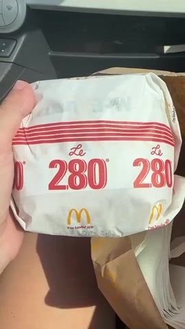 The video shows a person holding a white paper-wrapped McDonald's burger with red stripes and the number "280" printed on it. The burger is placed inside a brown paper bag along with several white napkins. The person's hand is visible reaching into the bag. The setting appears to be inside a car, with part of the car's interior visible in the background. The lighting suggests it is daytime.