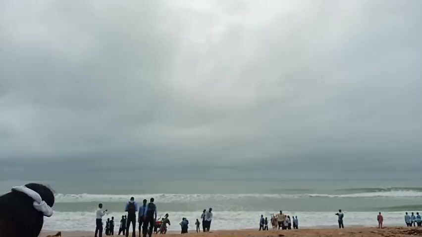 A person, presumably the video recorder, is standing on a beach with a view of other people and the ocean.

The sky is overcast with a thick layer of gray clouds. Below the clouds, the ocean stretches to the horizon with waves breaking near the shore. The beach is sandy, and several individuals are scattered along it, some standing in groups, others walking or interacting with what appears to be a horse. Most of the people seem to be wearing dark clothing. 

The person recording is partially visible in the bottom left corner, featuring the back of their head and a portion of their shoulder. They have dark hair pulled up with a light-colored scrunchie. The recording captures a scene of leisure and interaction on a cloudy day by the sea.