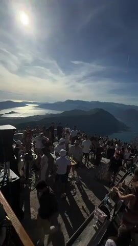 The video shows a wide-angle shot of a crowded outdoor gathering on a mountaintop overlooking a body of water and a mountainous landscape. The sun is visible and shining brightly in the upper left corner, casting long shadows. A large group of people, dressed in casual summer clothing, are mingling on a paved area. Some are standing, some are walking, and some appear to be interacting with each other. There is a railing in the foreground. The background features a scenic view of hills, mountains, and a bay or lake. The sky is blue with some scattered clouds. The overall impression is of a lively social event in a beautiful, elevated location.