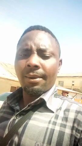 A man with short, dark hair, a mustache and a goatee is seen in a close-up shot, looking directly at the camera. He is wearing a grey and black checkered shirt. He is outdoors, as evidenced by the clear blue sky and buildings visible in the background. The buildings are mostly beige and have flat roofs. A sign for "KUME AFRICAN DELICACIES" is also visible in the background. The man appears to be talking.