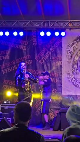 Two performers are on a stage, under various colored stage lights. A woman to the left plays a saxophone. A man stands to the right, singing into a microphone. Both wear black outfits. Behind them is a banner with the words "KYHOB SOUND" repeated, and another banner with a logo incorporating the words "THE RIDERS". The audience is visible in the foreground. The camera zooms in and out and changes focus.