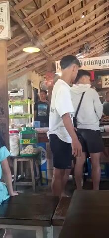 The video shows the interior of a food establishment with a thatched roof and wooden beams. In the foreground, a young person wearing a white t-shirt with text on the back and black shorts is standing, facing away from the camera. To their left, another young person in a light-colored hoodie and dark shorts is also visible. Further back, a man in a tan shirt and blue jeans is seen, as is another person in a red shirt. The establishment appears to be a casual eatery with tables and chairs. Various items, including food containers and what look like snacks, are displayed on shelves and counters. There are also ceiling fans providing air circulation. The overall atmosphere suggests a busy, everyday scene in a local food stall or restaurant.