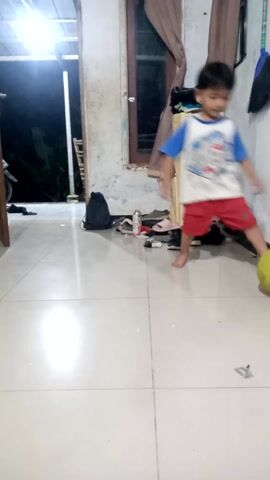 This media file shows two young children playing indoors. The setting appears to be a home, with tiled flooring and a doorway visible in the background. The lighting suggests it is nighttime or indoors with artificial lighting.

The first child, a boy wearing a blue and white t-shirt with red shorts, is holding a large yellow ball. He is positioned on the right side of the frame and appears to be interacting with the ball.

The second child, a girl wearing a black t-shirt and shorts, is on the left side of the frame. She is looking towards the camera and is holding a pair of black shoes. Her expression is somewhat unclear due to the image quality.

The children seem to be engaged in a playful activity, possibly a game involving the ball and the shoes. The overall mood seems casual and domestic. The scene is likely in Cibinong, Indonesia, as per the location context. There are no discernible weather conditions or specific time of day cues beyond the artificial lighting.