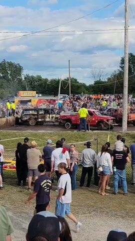 A demolition derby is underway with several heavily damaged vehicles in the arena. The crowd is seated in bleachers behind a safety barrier. Spectators are also standing in the foreground, watching the event. Several individuals in yellow safety vests appear to be workers or officials, and two people in firefighting gear are attending to the vehicles. One red pickup truck has the number 29 visible on its side. The sky is partly cloudy.