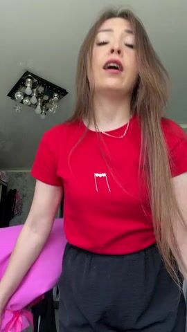 A young woman stands in a room, holding a large bouquet of pink lilies and roses. She is wearing a red t-shirt and black pants. The bouquet is wrapped in bright pink paper, creating a dramatic visual. The woman has long, brown hair and is looking at the camera with a playful expression. Her body is angled, and she is holding the bouquet in front of her. The room appears to be a domestic setting, with a patterned wallpaper and a ceiling light fixture visible. The overall mood is cheerful and celebratory, suggesting a gift or special occasion. There is no visible text in the video.