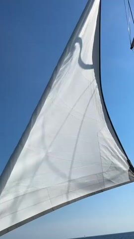 This media file features a close-up, low-angle shot of a large white sail on a sailboat against a clear blue sky. The sail is billowed by the wind, creating soft shadows and creases across its surface. The dark trim along the edges of the sail is visible, as is a hint of the sailboat's mast and rigging. In the distance, a faint, hazy line suggests the horizon where the sea meets the sky, indicating the sailboat is at sea. The bright sunlight suggests it is daytime, likely mid-morning or afternoon, given the intensity of the light and the clarity of the sky. The overall mood is serene and evokes a sense of freedom and adventure associated with sailing. There is no visible text in the media file. The location context provided is Ćunski, Croatia.