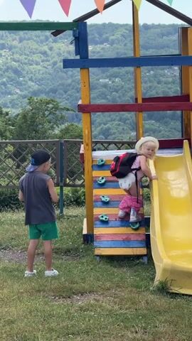 In a sunlit outdoor scene at a playground in Izmaylovka, Russia, a young boy wearing a dark grey sleeveless shirt, green shorts, and a blue baseball cap stands on a grassy area. He looks towards a colorful climbing wall attached to a yellow slide.

A young girl, wearing a light-colored hat, a pink shirt, a diaper, and pink pants, is climbing the wall. She has a red and black backpack on. Her legs are extended as she reaches for the next climbing hold, her shoes visible on her feet.

The climbing wall is constructed with wooden planks painted in alternating stripes of blue, yellow, and red. Bright green climbing holds are attached to the wall. The yellow slide curves downwards from the top of the structure.

Behind the playground, a lush, green forested hillside stretches upwards under a bright, overcast sky. Decorative triangular flags in various colors, including purple, red, orange, yellow, and green, are strung across the top of the playground structure, suggesting a festive atmosphere or celebration. The overall mood is one of playful outdoor activity, with the children engaged in climbing and playing.