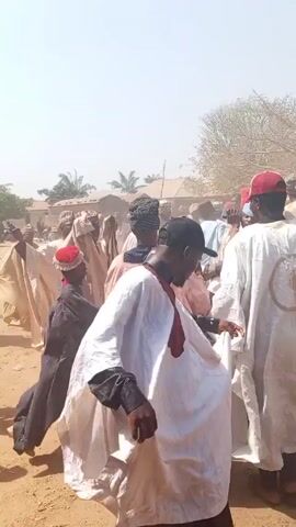 A crowd of people gather on a dirt lot under a bright sky. Most of the crowd is wearing traditional clothing, white robes and hats. Many of the crowd raise their arms. The environment appears rural, with houses and trees visible in the background. Dust is being kicked up from the ground. A man in a red shirt and hat stands in the foreground. A man in a white shirt with embroidered pattern also stands in the foreground and smiles.