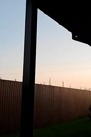 The video captures a view of a wooden fence from a covered porch. A dark, vertical pillar is in the foreground, obscuring part of the view. The sky is clear with a gradient of pale blue to light pink, suggesting sunrise or sunset. Some tall, thin plants are growing just above the fence. In the distance, faint power lines can be seen against the sky. To the right, part of a building's roofline is visible. The ground in front of the fence is covered in green grass. A gray utility box with a white door is mounted on the fence to the right of the pillar. A metal pole, possibly a utility pole, is also visible on the right, extending upwards into the sky.