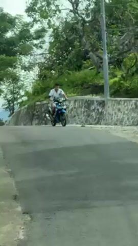 The video begins with a close-up view of a road surface with some indistinct writing or markings on it. The camera quickly shifts to show a man riding a blue scooter on a paved road. The road is bordered by a stone wall and lush green vegetation. The camera continues to move forward, providing a view of the road ahead, which is lined with trees and some buildings in the distance. The sky is overcast. The scooter rider continues down the road, becoming smaller as the camera continues moving forward.