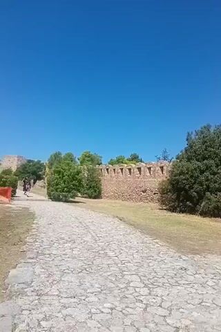 The video shows a person walking near a stone wall and a small building. The person, a woman wearing a white shirt and black backpack, is seen in the first few seconds from the side, walking along a cobblestone path. In the later part of the video, she is seen from the side again, holding a phone, and taking a picture of a flame. The environment appears to be outdoors, with a clear blue sky, green trees, and stone structures. The stone structures look like ancient ruins, possibly a castle or a fortress. The small building in the background looks like a church or a chapel, with a red door and windows. The path is made of cobblestones, and there is dry grass and some rocky areas around. The overall scene suggests a historical site or tourist attraction.