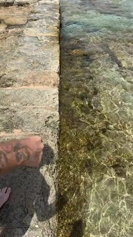 A person is walking along a stone ledge next to clear, shallow water. The person is wearing red swimming trunks and has tattoos on their left arm. The water is transparent, revealing rocks and some vegetation on the bottom. The sun is shining, casting shadows on the stone.