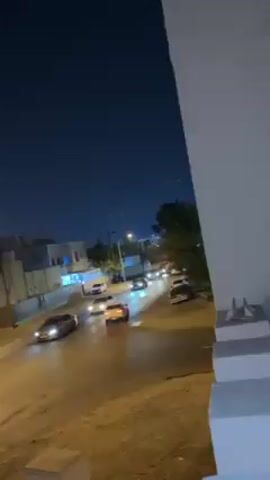 The video shows a street at night lined with buildings. Several cars are driving on the road, illuminated by streetlights. The sky is dark with faint lights visible in the distance. The camera pans upwards to capture a streetlight and the night sky before returning to the street scene.