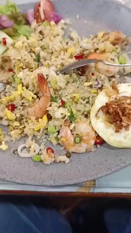 The video shows a close-up, overhead shot of a plate of fried rice. The plate is a dark gray color and contains a generous portion of fried rice, with several shrimp, pieces of squid, scrambled egg, chopped green onions, and red chili peppers mixed in. To the left of the rice, there is a small salad with lettuce, sliced cucumbers, and red onion. To the right of the rice, there is a fried egg topped with crispy fried onions. A fork and a spoon are resting in the rice. The food appears to be on a table, with a blurred background suggesting an indoor dining setting. The lighting is natural and seems to be coming from above. The video is shaky, suggesting it was filmed by someone holding the camera or phone.