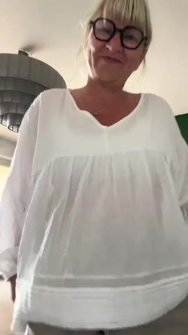 A woman with blonde hair and glasses, dressed in a white, flowing blouse and khaki-colored pants, stands in a well-lit living room. The room features a modern ceiling light fixture, a green painted wall with an artwork, and a grey couch. The woman adjusts her hair and smiles at the camera. A pink bag sits on the couch.