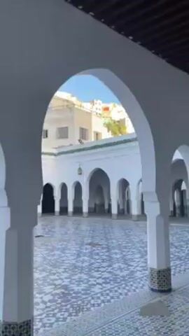 The video shows an outdoor courtyard with white arches and columns. The floor is tiled with blue and white geometric patterns. A wrought iron railing runs along the edge of a slightly elevated walkway. There are stairs leading down to the courtyard. In the background, buildings are visible. The video appears to be taken on a sunny day.