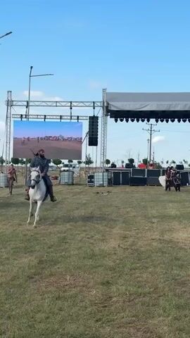 A reenactment is underway in a grassy field under a clear blue sky. A man on a white horse, dressed in historical garb, rides from left to right across the frame. In the background, a stage with a large screen and sound equipment is set up. Several other individuals in similar historical attire are present on the field, some interacting with each other and others with horses. Spectators are visible in the foreground, some holding up smartphones to record the event. The overall atmosphere suggests a historical demonstration or performance.