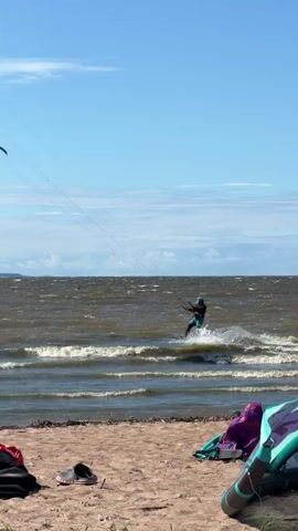 The video captures a bright, sunny day at a beach. The camera is positioned at eye-level, looking out towards the ocean. In the foreground, the sandy beach is visible, with a black bag and a pair of sandals lying on the sand. The ocean is choppy with waves, indicating a windy day. A person is kitesurfing in the distance, actively engaged in the sport, with spray of water visible around them. A kitesurfer's kite is also visible in the sky. The sky is blue with scattered white clouds. The overall impression is a vibrant and active outdoor scene.