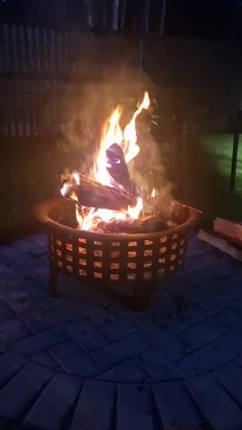 A fire burns in a metal fire pit on a blue brick patio at night. The fire pit has a decorative basket weave pattern. Logs are visible in the fire pit, with flames reaching upwards. Smoke rises from the fire. In the background, a wooden fence and trees are visible. To the right of the fire pit, there are stacks of firewood. The scene is dimly lit by the fire.