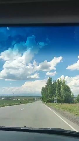 The video shows a view from inside a vehicle traveling on a road. The road is paved and has white lines marking the lanes. On the right side of the road, there are trees and a guardrail. In the distance, there are fields, houses, and mountains. The sky is blue with white clouds. A sign is visible near the trees on the right side of the road, but the text is illegible. The perspective suggests the viewer is in the front passenger seat or driving the vehicle.