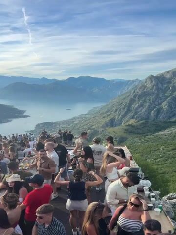 The video shows a crowded outdoor party or gathering on a mountaintop with a scenic view of a bay and mountains. Many people are standing, talking, and interacting, with some appearing to be dancing. There are tables set up with drinks and possibly food. The sun is setting, casting a warm glow over the landscape. The atmosphere appears festive and social.