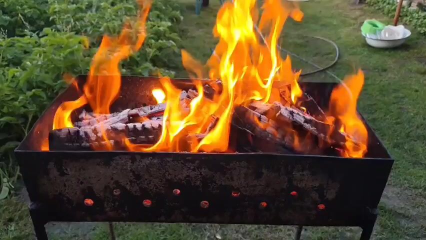 A close-up shot captures a metal barbecue grill with burning logs and bright orange flames. In the background, a green lawn surrounds the grill. Beyond the lawn, a light-colored house with multiple windows is visible, along with trees and a clear blue sky. A wooden shed or outbuilding with a corrugated metal roof is situated to the right of the house. The overall scene suggests an outdoor setting, possibly a backyard, during daylight hours.