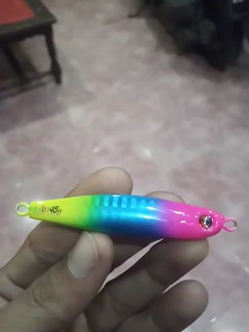 Here is a description of the video:

The video shows a person holding a brightly colored fishing lure in their hand. The lure is long and slender, with a rounded top and a pointed bottom. It is painted with a rainbow gradient, starting with pink at the top, transitioning to blue, and ending with green at the bottom. The lure has a small eye on the top and rings at both ends for attaching hooks. The brand name, "Klatri," and weight, "45g," are printed on the lure. The person's hand is holding the lure against a blurred background, which appears to be a tiled floor and some furniture.
