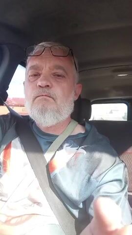 A middle-aged Caucasian man with gray hair and a gray beard wearing glasses and a gray t-shirt with a colorful design is sitting in the driver's seat of a vehicle. He is looking directly at the camera, then turns his head to the right, and then back to the camera. He raises his right hand and points his index finger towards the camera. The interior of the vehicle is visible, including a seatbelt. The exterior view through the windows shows buildings and trees. The lighting suggests it is daytime.