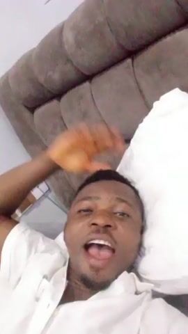 Here is a concise description of the video:

The video shows a man lying in bed, on a white pillow. He’s wearing a white shirt and has short, dark hair and a goatee. The man is looking at the camera and speaking. The headboard of the bed is upholstered in a gray, tufted fabric.