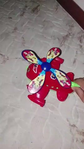 The video shows a red toy helicopter with a blue propeller. A hand is visible on the right side of the frame, holding a green object attached to the back of the helicopter. The helicopter is on a textured surface, likely a mattress or carpet. The propeller spins rapidly, blurring the images on its blades, which are decorated with colorful cartoon designs. The helicopter also appears to be spinning. The lighting is dim.