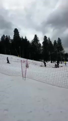 The video shows a ski park on a cloudy day. The ground is covered in snow and there are several people skiing and snowboarding. In the foreground, there is a safety net. Behind the net, there is a small jump and a large inflatable landing pad. There are red flags with white lettering visible. Trees are visible in the background.