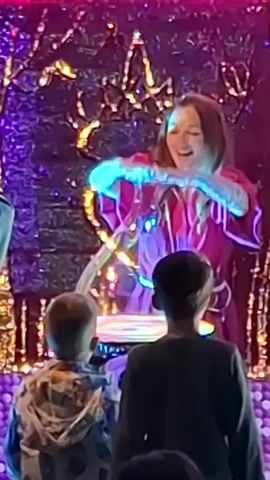A female performer in a red outfit with puffy sleeves stands behind a rotating light display, smiling and gesturing. Two children are standing in front of her, watching the performance. The background features a sparkly, illuminated decoration, and there are additional lights changing color throughout the scene.