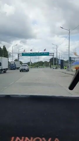 The video shows a wide road leading under a green archway that reads "WELCOME TO THE CITY OF IMUS". The archway is adorned with multiple Philippine flags. There are streetlights and some foliage visible on either side of the road. A white pickup truck is seen driving towards the archway on the left lane. Further down the road, several people are visible, and on the right side, there appear to be some structures and more people. The sky is cloudy. The camera appears to be positioned inside a vehicle, looking out through the windshield.