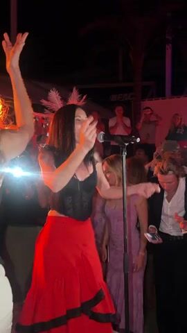 A woman with long dark hair, wearing a black sleeveless top and a red tiered skirt with black trim, sings into a microphone on a stand, moving her arms and body. She is illuminated by bright, warm light sources, one of which appears to be a phone flashlight held by a person in a white dress to her left, whose arm is raised. To the singer's right, a woman in a light purple dress smiles and dances, next to a man in a white shirt and black jacket. Several other people are visible in the darker background, watching the performance in an outdoor night setting.