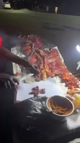 At night, a person cuts pieces of cooked meat on a table covered with aluminum foil and parchment paper. To the right is a white bowl containing a dark orange sauce. A second person shines a light on the meat. More cooked meat is arranged in rows on skewers to the right side of the table. The scene is dimly lit by street lights, other food stands, and the handheld light.