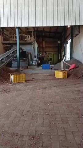 The video depicts the interior of a large, open industrial space. The floor is made of red bricks and scattered with debris. In the foreground, there are two yellow crates. Further into the space, there is an elevated platform with a conveyor belt that leads down to a pile of material. A large machine, possibly a processing unit, is visible near the conveyor belt. A person is standing in the middle distance, near a blue crate. The space is illuminated by overhead lights.