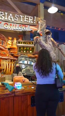 The video displays a shooting gallery attraction with a dinosaur and wild west theme. In the foreground, a person with long, curly brown hair wearing a light blue shirt and dark pants stands with their back to the camera, looking at the attraction. To their right, a person wearing a black t-shirt and brown pants, with a black backpack with the word "RACE" visible on it, also stands with their back to the camera, looking at the attraction. Another person, partially visible on the left, is also looking at the attraction. The attraction features a rustic wooden structure with a sign that reads "BONE BLASTIN' SHOOTIN' GALLERY." Behind the sign, there are various props including faux rock formations, dinosaur bones, and targets. A camouflaged jeep-like vehicle with cartoon eyes is positioned within the attraction. There are also several glowing buttons or screens on the counter in front of the attraction, where the people are standing. The overall environment appears to be indoors, likely an amusement park or arcade, with overhead lighting visible.